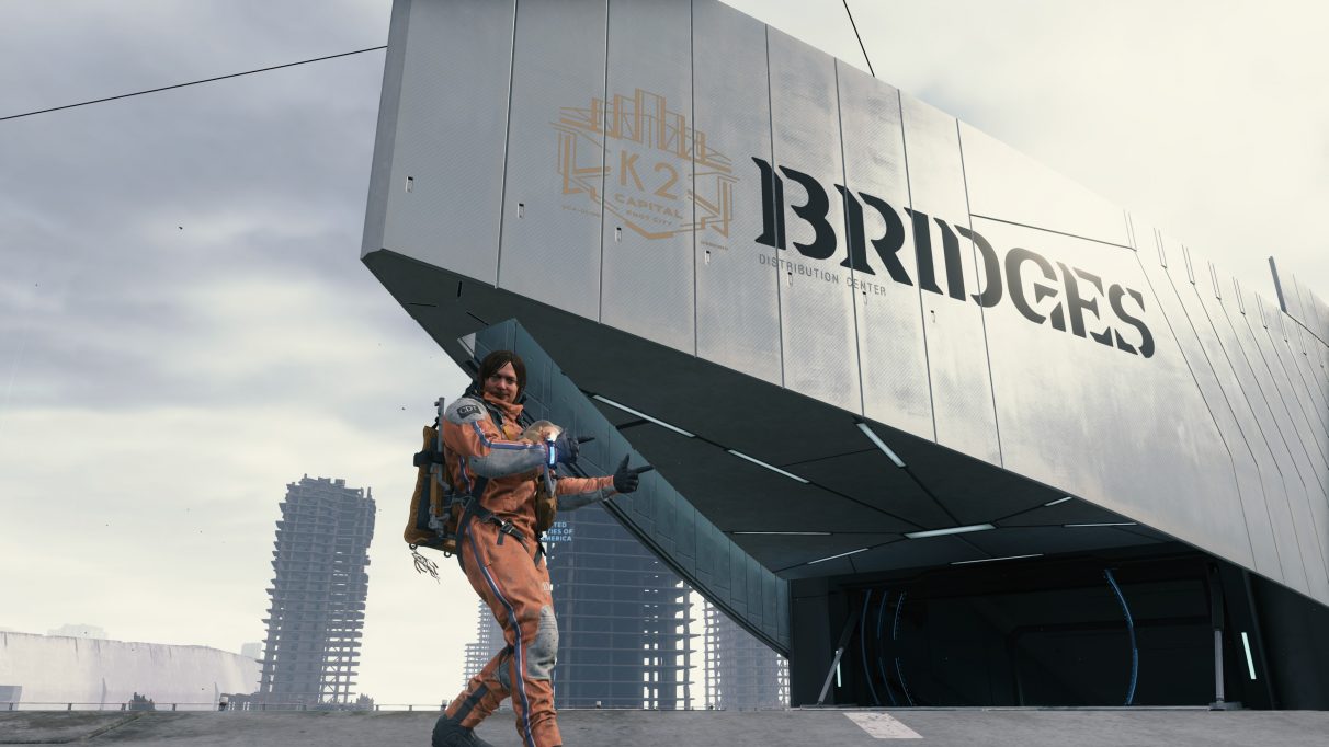 A screenshot of Sam Bridges doing a thumbs up sign outside a Bridges depot facility in Death Stranding.