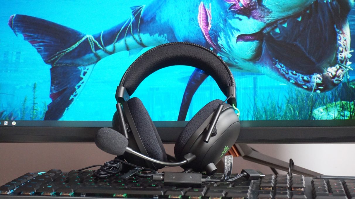 A photo of the Razer Blackshark V2 gaming headset perched on top of a keyboard and resting against a monitor with a shark wallpaper.