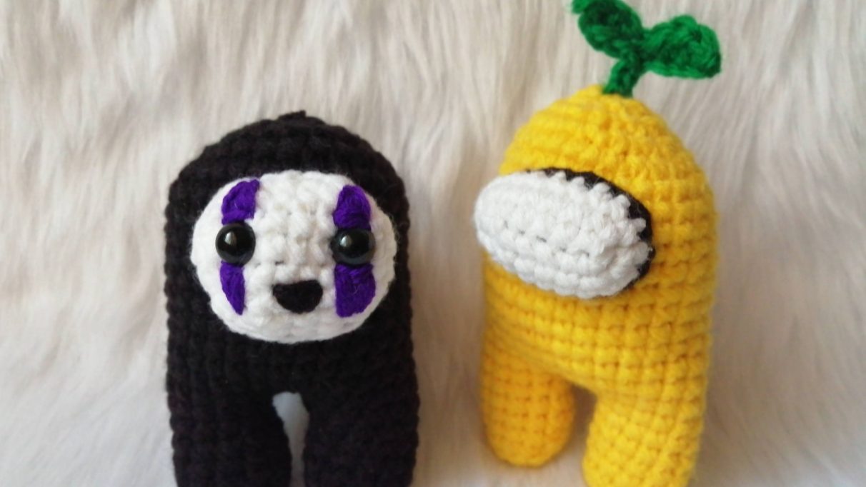 A picture of two teeny crewmate amigurumi - stuffed crocheted figures - of Among Us crewmates made by Reddit user prudence0381. One is a yellow crewmate with a little seedling sprouting out of the top of its head. The other is black, and resembles No Face from Spirited away, although it looks very happy.